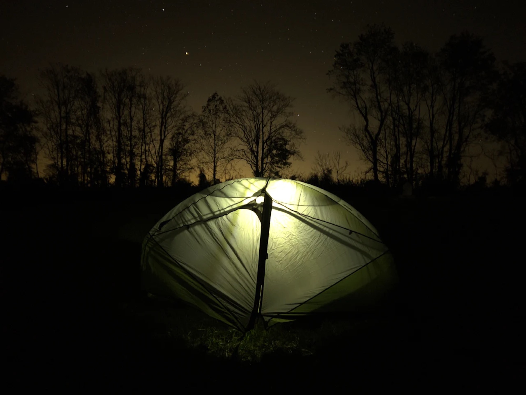 A tent glowing in the dark with stars visible in the sky above silhouetted trees