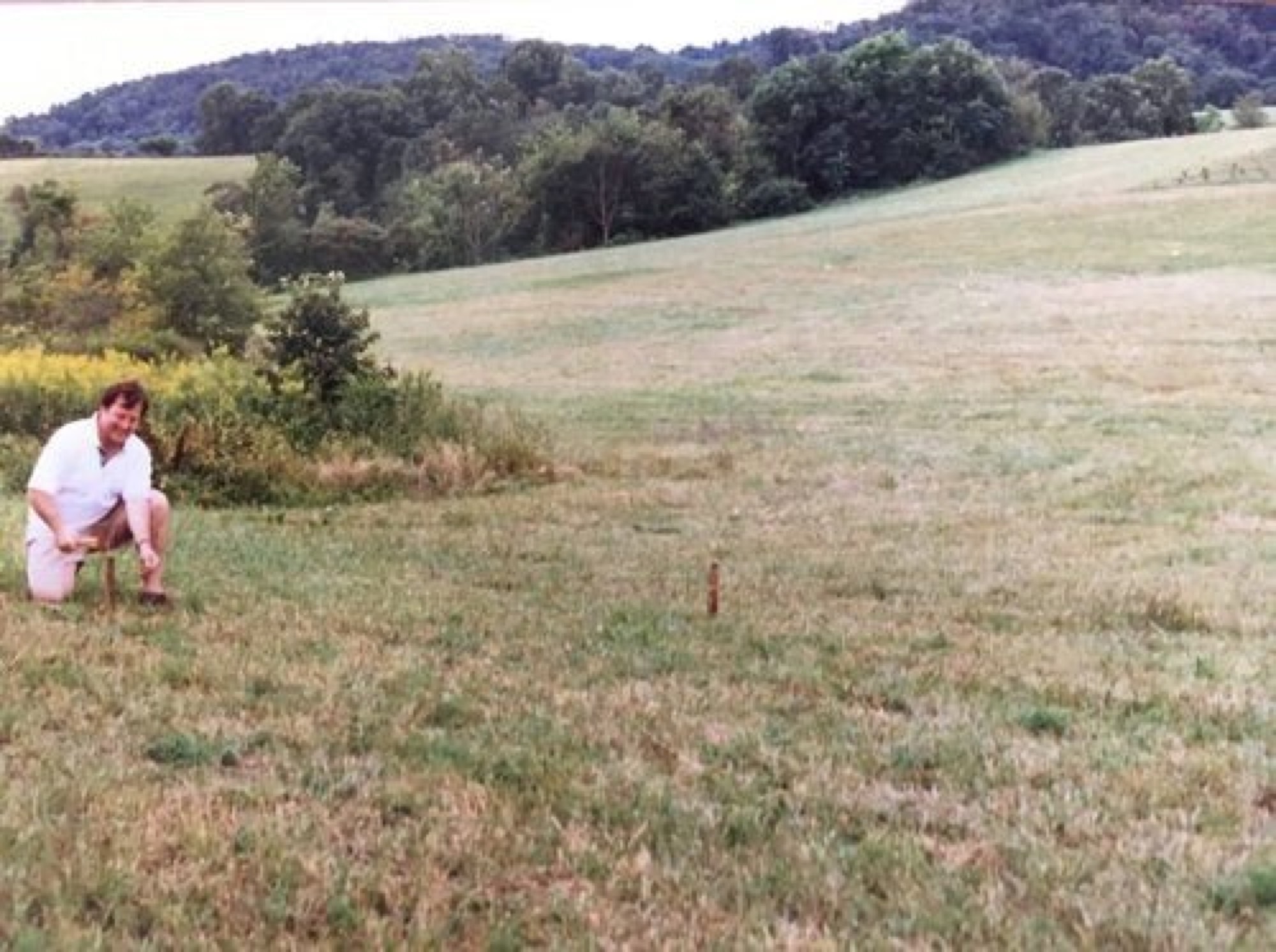 A woman crouched in an empty hillside field beside a single survey stake, before the vineyard was planted