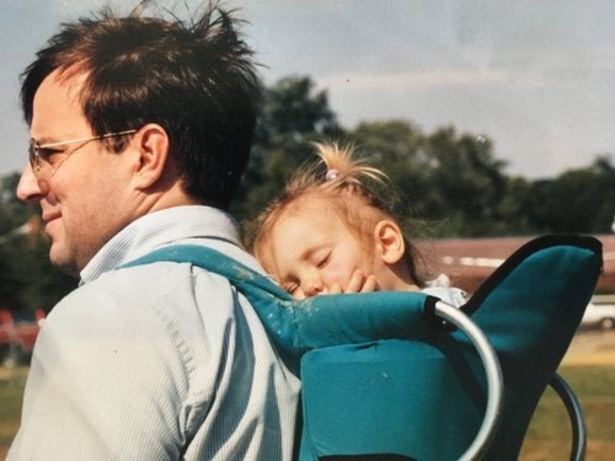 A man carrying a sleeping toddler in a backpack carrier, walking through open grass
